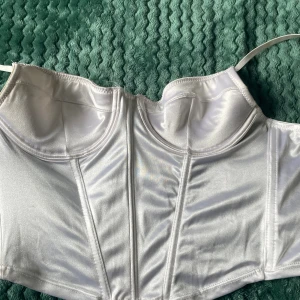Vit satin korsett med hakknäppning - Säljer en vit korsett i glansigt satinmaterial med bygel och formade kupor. Toppen har justerbara axelband och hakknäppning baktill för perfekt passform. Snygga sömmar framtill ger en cool och strukturerad look. Perfekt för att lyfta din outfit!