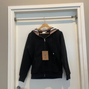 Svart Burberry hoodie med dragkedja - Svart hoodie från Burberry i storlek M med klassisk rutig huva i beige, rött och svart. Tröjan har dragkedja framtill, snörning vid huvan och en stor ficka på magen. Perfekt för dig som gillar stilrena och lyxiga streetwear-plagg.