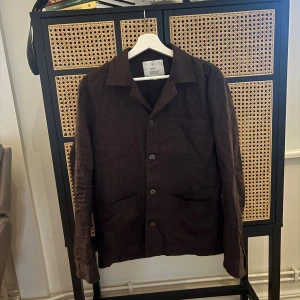 Brun overshirt från Bläck - Stilren brun overshirt från Bläck med klassisk krage och knäppning framtill. Jackan har tre fickor på framsidan och är tillverkad i ett mjukt bomullsmaterial. Perfekt för lager-på-lager och enkel att matcha med olika outfits.