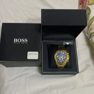 Guldig klocka från Hugo Boss - Säljer en snygg klocka från Hugo Boss med guldig länk och blå urtavla. Klockan har flera små urtavlor och detaljer i guld, samt tachymeter runt boetten. Kommer i originalask, perfekt för dig som gillar stilrena accessoarer.