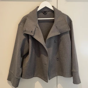 Grå kapp från Gina Tricot - Säljer en snygg grå kappa från Gina Tricot i storlek S. Jackan har bred krage, dubbelknäppning med stora knappar och långärmade ärmar. Materialet är mjukt och känns som ullblandning, perfekt för kyliga dagar. nypris omkring 500 kr