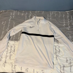Vit Nike half zip träningströja S - Vit långärmad träningströja från Nike med svart logga och svart rand över bröstet. Tröjan har half zip och står för en sportig look. Tillverkad i Dri-Fit polyester som andas och passar perfekt till träning.
