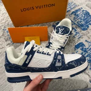 Louis Vuitton Trainer Denim Blue White - Originala Louis Vuitton sneakers i vitt läder och blå denim med monogrammotiv. En ikonisk modell med exklusiv och sofistikerad design. ✅ Skick: Nya, aldrig använda. 📏 Storlek: 42 📦 Snabb och noggrann frakt Levereras med originallådan från Louis Vuitton. Perfekt för dig som söker en lyxig men ändå streetwear-stil.