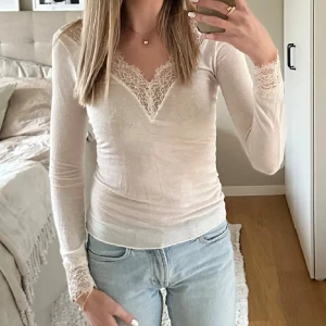 Intimissmi Vit långärmad spetsblus med v-ringning - Supersnygg vit blus med lång ärm och v-ringad hals, dekorerad med spetsdetaljer både vid halsringningen och ärmsluten. Modellen är slim och sitter tajt mot kroppen, perfekt för dig som gillar en feminin och stilren look. Materialet känns mjukt och stretchigt.