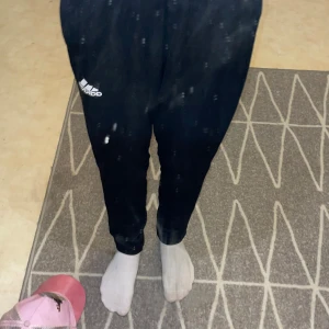 Svarta Adidas mjukisbyxor i storlek S - Svarta mjukisbyxor från Adidas med vit logga på låret. Byxorna har smal passform, resår i midjan och vid benslut samt dragkedja vid fickorna. Tillverkade i mjukt och bekvämt polyestermaterial, perfekta för chill eller träning.