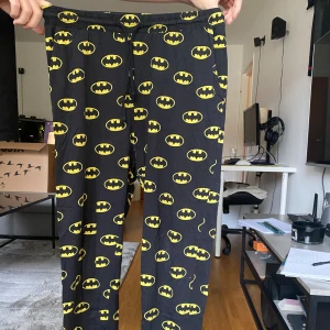 Svarta Batman-mjukisbyxor med tryck - Svarta mjukisbyxor med elastisk midja och snörning, täckta av gula Batman-loggor över hela tyget. Perfekta för chill dagar hemma eller när du vill visa din kärlek för superhjältar. Mjukt och bekvämt material.