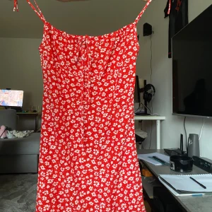 Röd blommig kortklänning med smala axelband - Superfin röd klänning med vita blommor och smala axelband som knyts upptill. Modellen är kort och har en lätt utställd form som ger en skön och avslappnad vibe. Perfekt för varma dagar och har ett somrigt mönster som sticker ut.