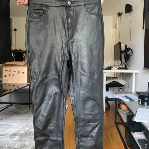 Svarta byxor i skinnimitation - Säljer ett par svarta byxor i skinnimitation med klassisk femficksdesign och knapp- samt dragkedjestängning. Byxorna har raka ben och en stilren look som passar till många olika outfits. Perfekta för dig som vill ha en edgy vibe.