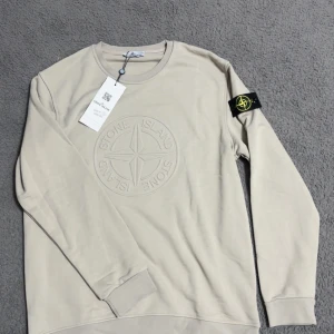 Beige Stone Island sweatshirt - Snygg beige sweatshirt från Stone Island med stort broderat logomärke på bröstet och klassisk patch på ärmen. Tröjan har rund halsringning och långa ärmar, perfekt för en clean och stilren look.
