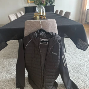 Svart pufferjacka från McKenzie M - Svart pufferjacka från McKenzie med huva och dragkedja. Jackan har quiltad framsida och rygg, släta ärmar med reflexdetaljer och en ficka med dragkedja på ena ärmen. Perfekt för kyliga dagar och snygg streetstil.