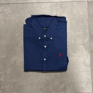Blå skjorta från Ralph Lauren - Snygg marinblå skjorta från Ralph Lauren med klassisk krage och vita knappar framtill. På bröstet finns den ikoniska röda Polo-loggan broderad. Skjortan är långärmad och tillverkad i mjuk bomull, perfekt för en clean och stilren look. Passar mer som S än M enligt mig