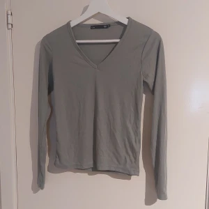Grå ribbad långärmad topp 157 - En enkel och stilren grön långärmad topp från 157 i storlek XS. Toppen är ribbad med v-ringning och har en smal passform. Tröjan har ett litet "hål" eller vad man ska kallade (se bild). 