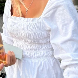 Vit omlottklänning med puffärm - Säljer en vit klänning med puffiga långärmar och smockad midja. Klänningen har fyrkantig ringning och är tillverkad i mjukt bomullstyg. Perfekt för dig som gillar romantisk och trendig stil med volym och detaljer.