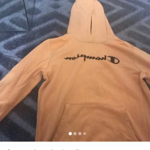 Beige hoodie från Champion XL - Säljer en beige hoodie från Champion i storlek XL. Tröjan har svart logga framtill, huva och en stor magficka. Perfekt för dig som gillar en chill och avslappnad stil. Materialet är mjukt och skönt, troligtvis bomull eller polyesterblandning.