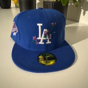 Blå LA Dodgers 59FIFTY keps med broderi - Snygg blå LA Dodgers 59FIFTY keps från New Era med broderade vita LA-loggor och lila blommor på framsidan. På sidan finns ett lila 1988 World Series-märke. Undersidan av skärmen är ljuslila. Perfekt för dig som gillar baseball och streetwear.