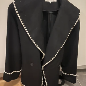Svart kappa med vit kant från Bubbleroom - Stilren svart kappa från Bubbleroom i storlek S med bred krage och dekorativa vita kanter längs krage, ärmslut och nederkant. Kappan har knäppning framtill och en klassisk, rak passform som ger en snygg siluett.