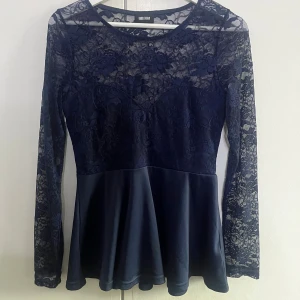 Marinblå spetsblus med peplum - Supersnygg mörkblå blus från Bubble Room med transparent spets upptill och på ärmarna. Blusen har lång ärm, rund halsringning och en peplum-nederdel i slätt tyg. Supersnygg till festliga sammanhang!