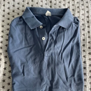 ARKET pikétröja i bomul, storlek M - Snygg blå pikétröja i klassisk modell med krage och tre vita knappar framtill. Tillverkad i mjuk bomull som känns skön mot huden. Perfekt för en chill och stilren look. Kortärmad och passar till många olika outfits. I nytt skick!