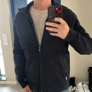 Svart cardigan jacka från Massimo Dutti - Svart jacka med snygga stickade ärmar och hel dragkedja framtill. Jackan har två fickor med dragkedja och en clean, modern look. 