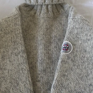 Grå stickad polotröja från Devold XS - Supermysig gråmelerad polotröja från Devold i grovstickad ull. Tröjan har hög polokrage, ribbade muddar och en snygg patch med norska flaggan på bröstet. Perfekt för kalla dagar och riktigt skön att ha på sig.