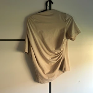 Beige t-shirt med snörningsdetalj - Snygg beige t-shirt med korta ärmar och cool snörningsdetalj på sidan som ger en unik twist. T-shirten har en avslappnad passform och är gjord i mjukt bomullsmaterial. Perfekt för dig som gillar stilrena plagg med en liten extra detalj.
