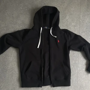 Svart hoodie från Polo Ralph Lauren - Svart hoodie från Polo Ralph Lauren med vit snörning i huvan och röd broderad logga på bröstet. Tröjan har dragkedja, kängurufickor och är tillverkad i mjuk bomullsmix. Perfekt för en chill och stilren look.