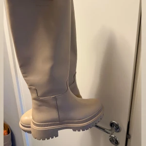 Beige höga boots  - Snygga beige boots med högt skaft och chunky platt sula. Tillverkade i slätt syntetmaterial med minimalistisk design och rund tå. Perfekta för att lyfta din höst- eller vinterstil med en modern touch.