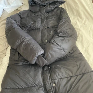Svart lång pufferjacka med huva - Säljer en svart lång pufferjacka med huva och snygga detaljer som tryckknappar och dragkedja. Jackan har en quiltad look och är perfekt för kalla dagar. Märke med logga på ärmen ger extra stilpoäng. Riktigt skön och varm att bära. vinter jacka storlek M. frågor? skriv