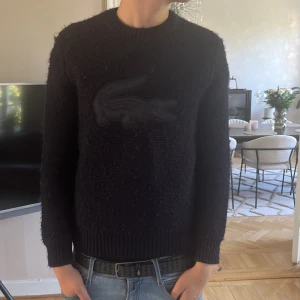 Svart stickad tröja från Lacoste - Säljer en svart stickad tröja från Lacoste med klassisk passform. Tröjan har ett stort ton-i-ton Lacoste-krokodilmärke på bröstet, ribbade muddar och rund hals. Perfekt för dig som gillar stilrena och varma plagg med en sportig touch.