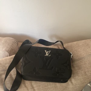 Svart Louis Vuitton axelväska i skinn - Säljer en svart axelväska från Louis Vuitton i skinn med präglat monogrammönster och silverfärgad LV-logga framtill. Väskan har justerbar axelrem och dragkedja upptill. Perfekt storlek för det viktigaste och riktigt snygg att bära crossbody.