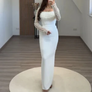 Vit långklänning med pärldetaljer - Elegant vit långklänning med figurnära passform och transparenta långärmar dekorerade med pärlor. Klänningen har fyrkantig ringning och pärlbroderier över bysten och ärmarna, vilket ger en lyxig känsla. Perfekt för speciella tillfällen. Använd enbart en gång.