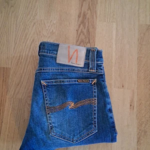 Blå Nudie jeans - Snygga blå jeans från Nudie Jeans med klassisk femficksdesign och orangea kontrastsömmar. Jeansen har en rak passform och är tillverkade i slitstarkt denimtyg av bomull. Perfekta för dig som gillar en tidlös och clean look. 27/32