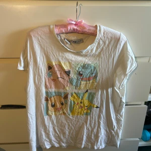 Vit Pokémon t-shirt från FB Sister - Vit t-shirt från FB Sister med Pokémon-tryck framtill som visar Pikachu, Eevee, Jigglypuff och Squirtle i färgglada rutor. Klassisk rund hals och korta ärmar. Perfekt för dig som älskar Pokémon och vill ha en chill och lekfull stil.