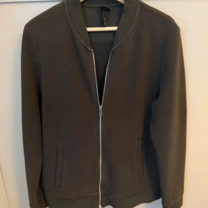 Svart bomberjacka med dragkedja - Säljer en stilren svart enkeljacka med silverfärgad dragkedja framtill. Jackan har ribbade muddar vid krage och ärmslut samt två fickor på sidorna. Perfekt för dig som gillar en clean och enkel look. Priset går alltid och diskutera hör av dig om du har flera frågor 