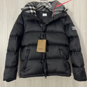 Svart pufferjacka från Burberry - Svart pufferjacka från Burberry med klassiskt rutigt foder i huvan. Jackan har dragkedja och knappar framtill, två fickor med dragkedja och logga på ärmen. Perfekt för kalla dagar och riktigt snygg med den ikoniska Burberry-looken.