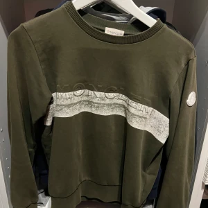 Olivgrön Moncler sweatshirt med logga - Snygg olivgrön sweatshirt från Moncler med vit logga på ärmen och en bred, vit rand över bröstet. Tröjan har rund hals, ribbade muddar och en clean look med diskret Moncler-tryck framtill. Perfekt för dig som gillar streetwear vibes.