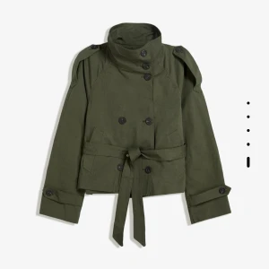Olivgrön kort trenchcoat med bälte - Snygg olivgrön trenchcoat med hög krage, dubbelknäppning och axelklaffar. Jackan är croppad och har bälte i midjan samt knappar vid ärmslut. Perfekt för dig som gillar en stilren och edgy look.