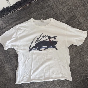 Vit t-shirt med pantertryck - Vit t-shirt med coolt tryck av en svart panter och en person med röd scarf på framsidan. T-shirten har rund hals och korta ärmar, tillverkad i mjuk bomull. Passformen är normal och trycket ger en unik vibe till din outfit.