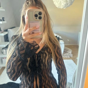 Svart spetsblus med knyt från Pieces - Säljer en svart transparent spetsblus från Pieces i storlek XS. Blusen har långa ärmar, v-ringning och snygg knytning framtill. Mönstret är vågigt med spets. Perfekt att styla lager på lager eller över ett linne.