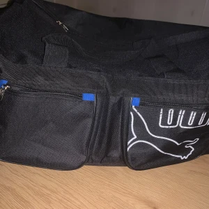 Svart träningsväska från Puma - Stor svart träningsväska från Puma med klassisk logga i vitt på framsidan. Väskan har två ytterfickor med dragkedja och blå detaljer, samt rejäla handtag. Tillverkad i slitstark polyester och perfekt för gymmet eller sportaktiviteter.