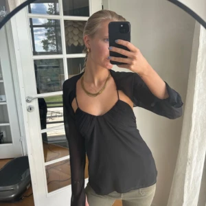 Svart omlottopp med öppen rygg - Säljer en svart omlottopp med tunna axelband och långa, transparenta ärmar. Toppen har öppen rygg med knyt och en snygg drapering framtill. Perfekt för dig som gillar trendiga och unika detaljer.