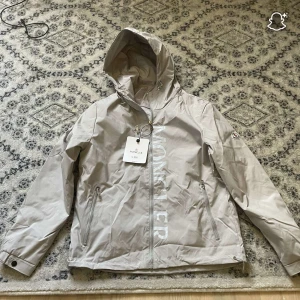 Beige vindjacka från Moncler med huva - Snygg beige vindjacka från Moncler med stor logga framtill, huva och dragkedja. Jackan har två fickor med dragkedja, justerbar nederkant med snörning och klassisk Moncler-patch på ärmen. Perfekt för dig som vill ha en clean och stilren look.