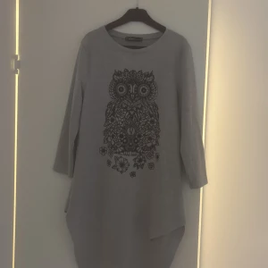 Grå klänning med uggletryck FB Sister - Säljer en grå långklänning från FB Sister i storlek XS. Klänningen har ett stort svart uggletryck med blommor framtill, rund hals och trekvartsärm. Materialet är mjukt och känns som bomull. Asymmetrisk nederkant ger en cool vibe.