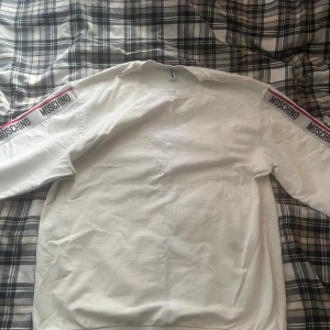 Moschino  - Vit sweatshirt från Moschino med svarta logoband längs ärmarna och en röd linje som detalj. Tröjan har rund halsringning, långa ärmar och ribbade muddar. Den har en flack på ena armbågen som ni kan se på sista bilden