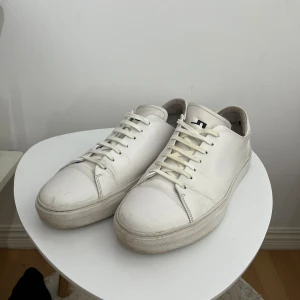Vita  sneakers - J.lindeberg - Stilrena vita sneakers med rund tå och platt sula. Skorna har snörning och är tillverkade i skinn med diskret svart logga på plösen. Perfekta för en clean och modern look. Från J lindeberg