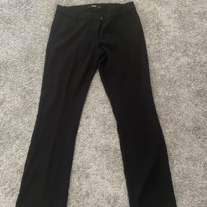 Svarta kostymbyxor från Bik Bok - Klassiska svarta kostymbyxor från Bik Bok med raka ben som är lätt utsvängda nertill och medellåg midja. Byxorna har bälteshällor, knapp och dragkedja framtill. Perfekta för en clean och stilren look. Materialet är mjukt och följsamt för extra komfort.