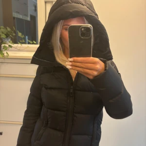 Svart pufferjacka med huva - Säljer en svart pufferjacka med stor huva och dragkedja framtill. Jackan är vadderad och har en klassisk stil med kvinnlig passform som formar fint. Passar dig som vill ha en stilren och enkel jacka som är trendig.