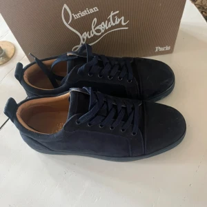 Mörkblå sneakers från Christian Louboutin - Snygga mörkblå sneakers från Christian Louboutin i mocka med klassisk röd sula och diskret logga undertill. Skorna har rund tå, platt sula och snörning. Insidan är fodrad i beige skinn för extra komfort. Perfekta för dig som vill sticka ut med exklusiv stil.