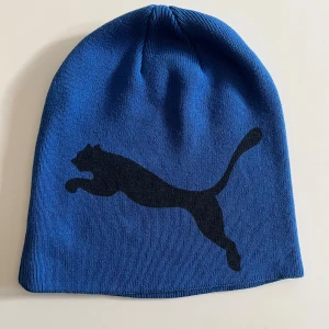 Blå mössa från Puma med logga - Snygg blå mössa från Puma med stor svart logga på framsidan och liten Puma-text bak. Tillverkad i stickad polyester, perfekt för kalla dagar. Enkel och stilren design som passar till det mesta.