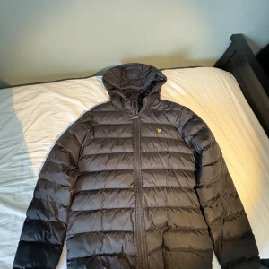 Svart dunjacka med huva från Lyle & Scott - Säljer en svart dunjacka från Lyle & Scott med huva och dragkedja. Jackan har det klassiska guldiga logomärket på bröstet och är quiltad med horisontella sömmar. Perfekt för kalla dagar och riktigt skön att bära. Hål i armhålan men om man kan sy så löser man.